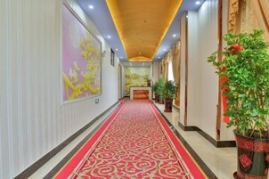 Corredor de hotel com carpete vermelho, balcão de recepção e plantas em vasos.