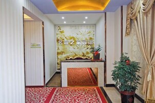 Un área de recepción con una pared decorativa, un mostrador de recepción, una alfombra roja estampada y una planta en maceta.