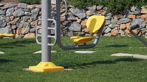 An outdoor exercise station with yellow and gray components, set against a backdrop of a stone wall and greenery. - Small Villa Emy Exclusive (Sporades)