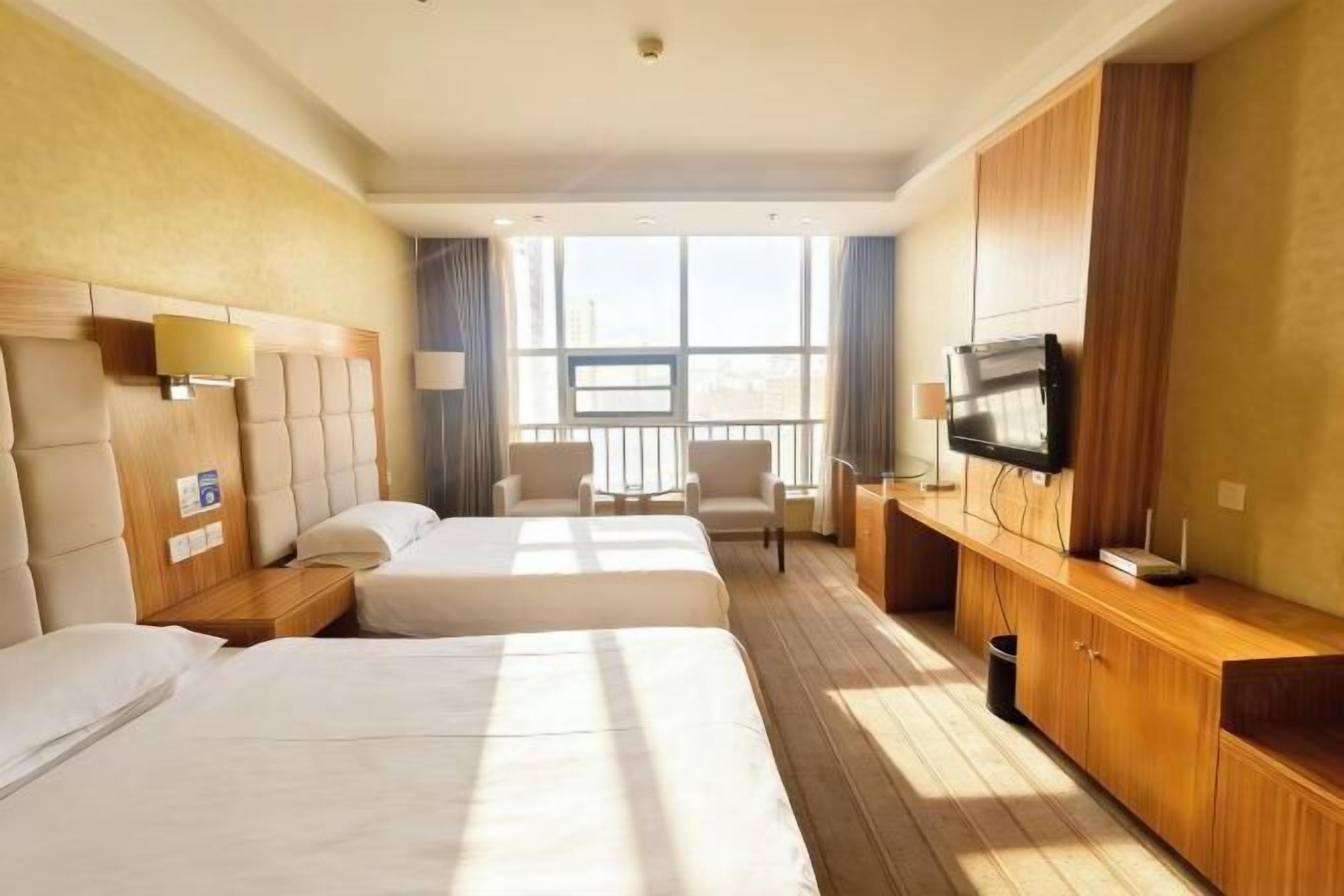 A hotel room with two beds, a TV, a desk, and a window with a city view.