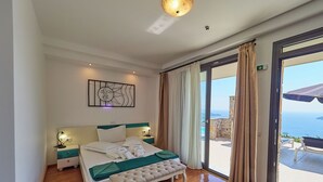 Habitación de hotel con cama, mesitas de noche, un banco y un amplio ventanal con vista al mar.