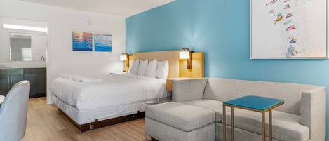 A hotel room with a bed, a grey sofa, a small table, and a wall with artwork.