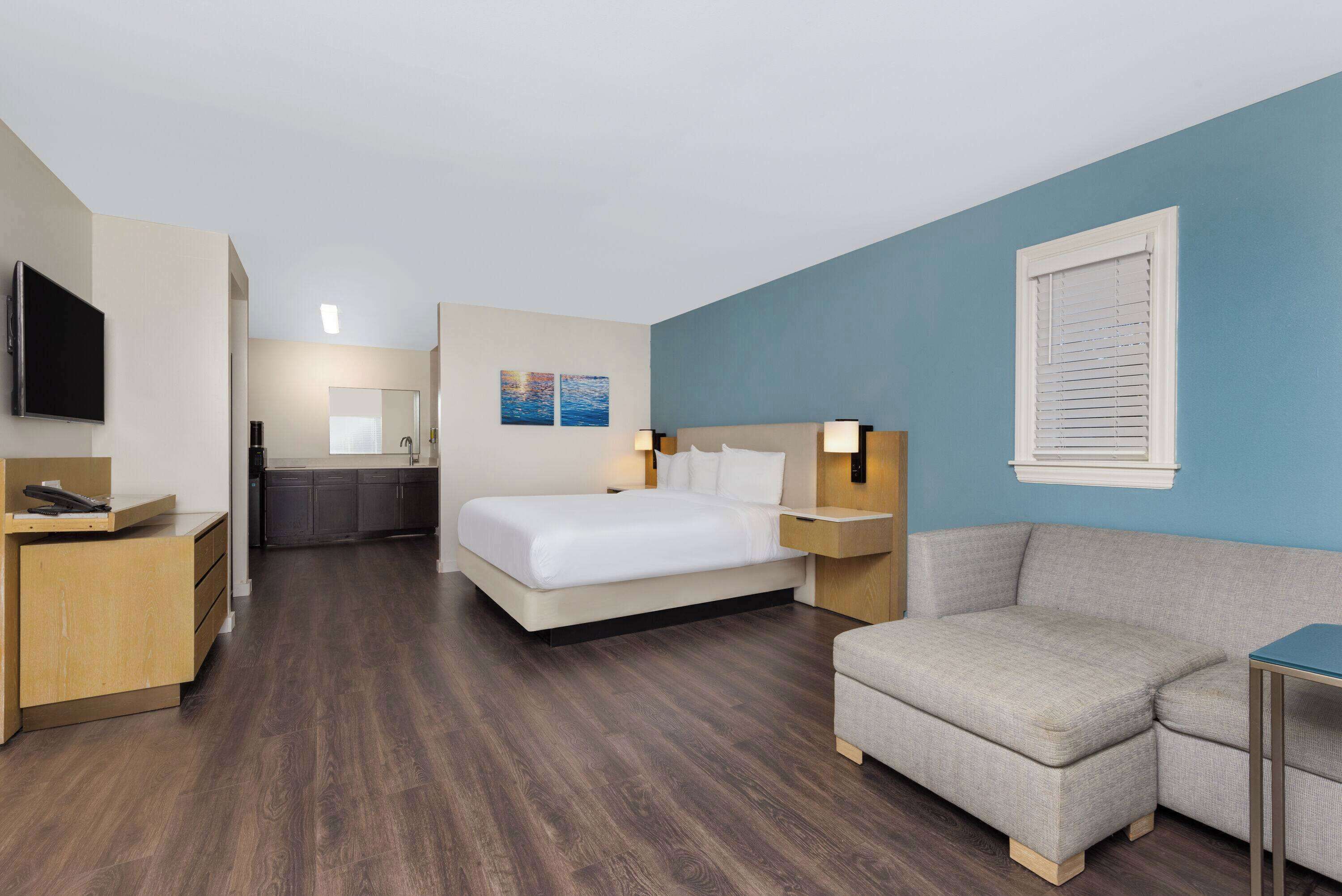 A hotel room with a bed, a sofa, a desk, a TV, and a bathroom visible in the background.