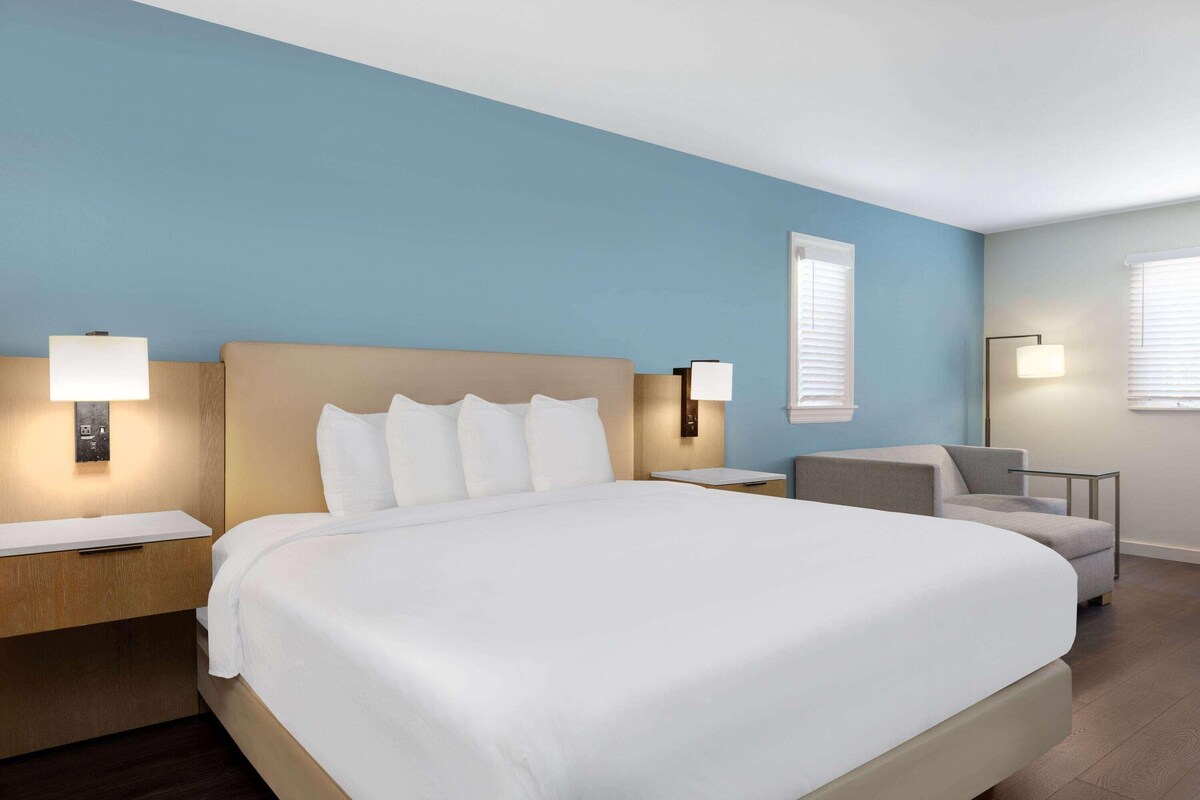 Una habitación de hotel con una cama grande, mesitas de noche, una silla y dos lámparas fijadas a la pared.