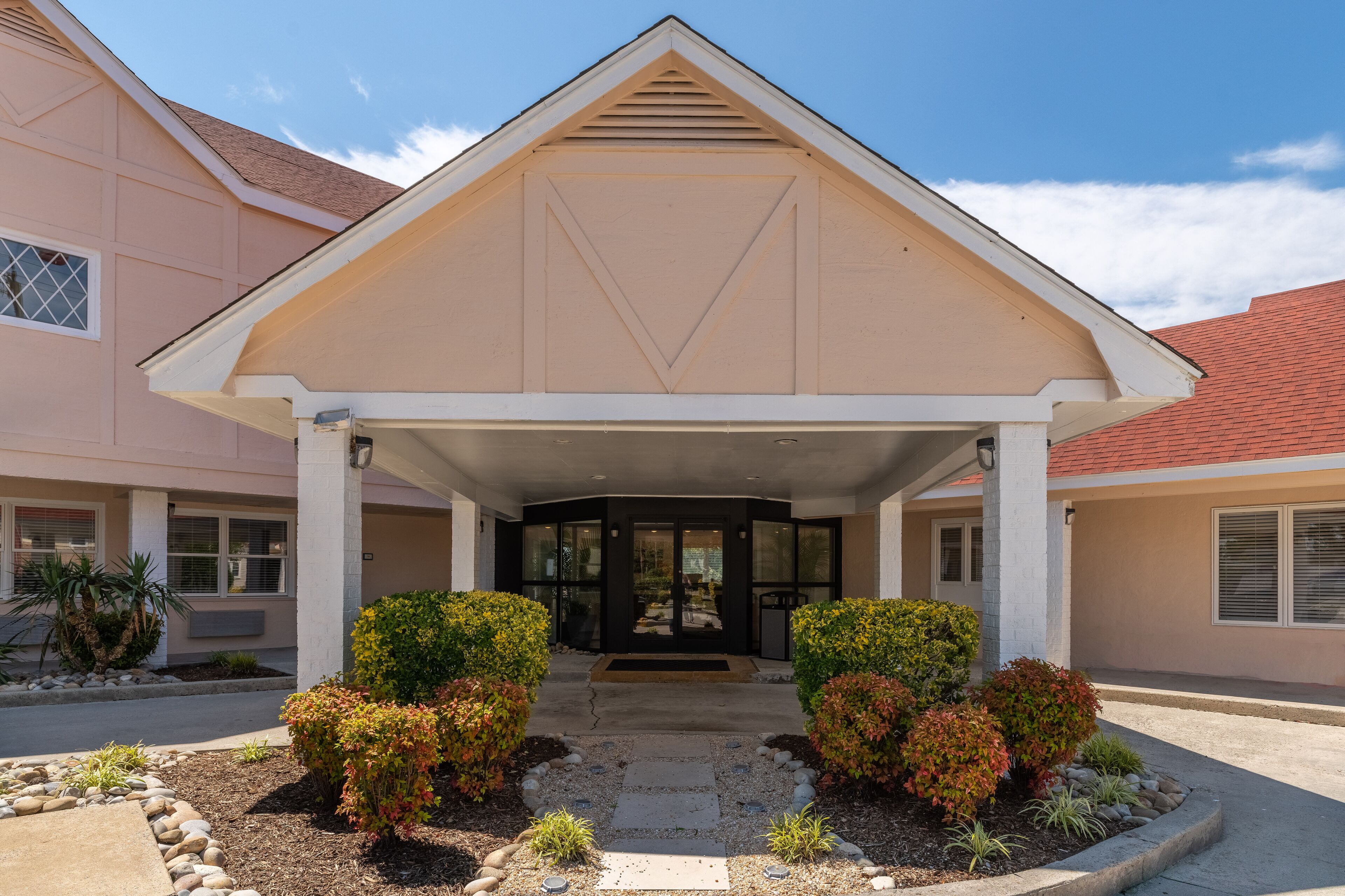 A building with a covered entrance, a triangular roof, and a landscaped front yard with shrubs and small plants.