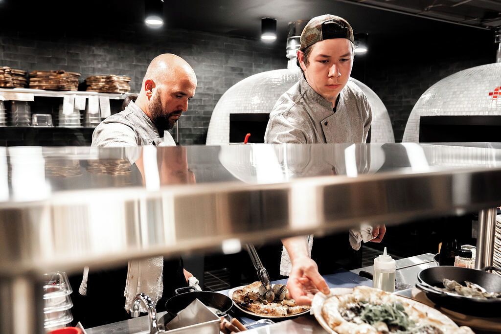 A kitchen with two chefs preparing food, a stainless steel counter, and a brick wall in the background.