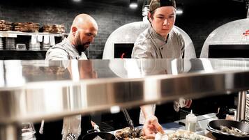 A kitchen with two chefs preparing food, a stainless steel counter, and a brick wall in the background.