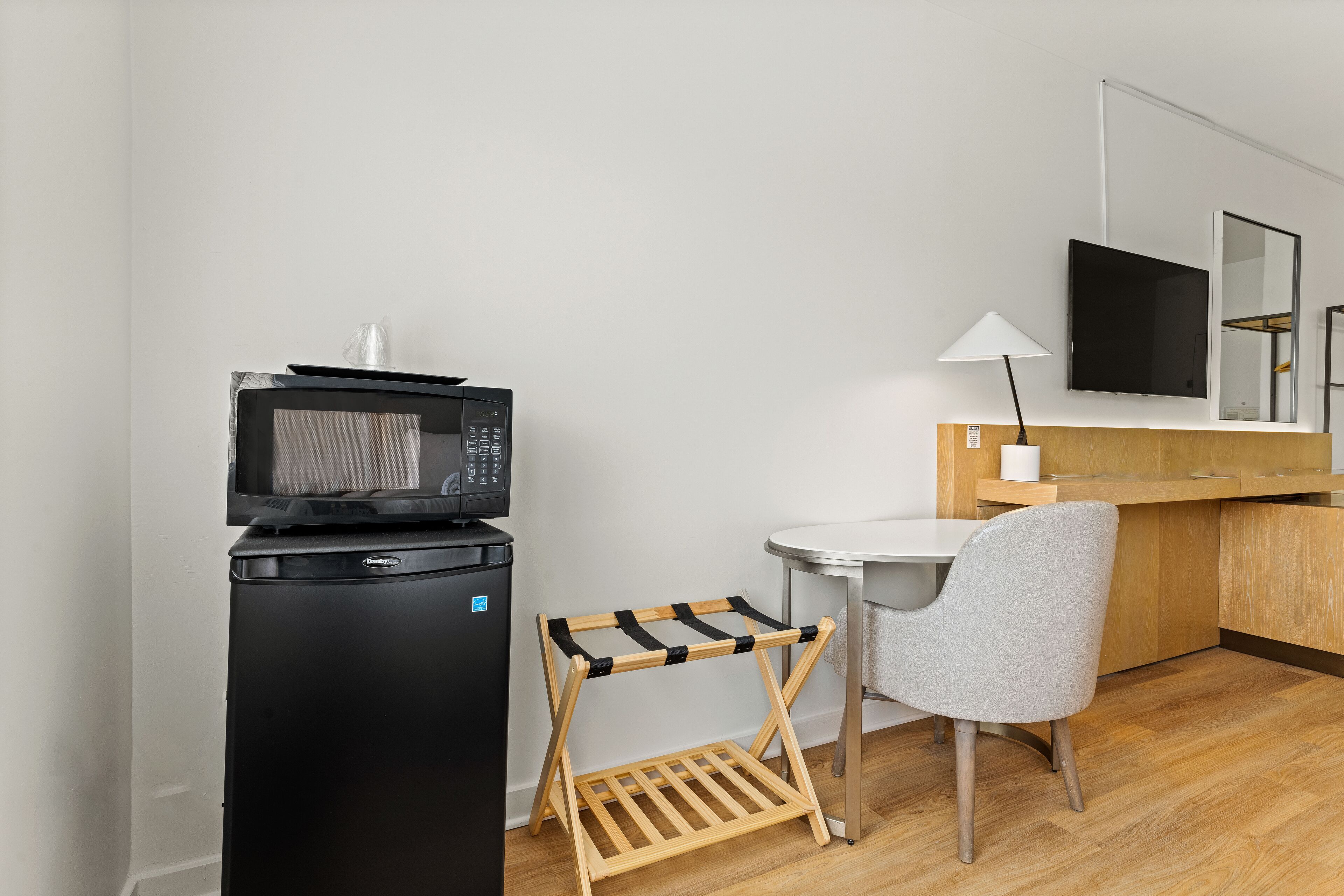 A compact kitchenette with a microwave, a small refrigerator, a wooden drying rack, a chair, and a table with a lamp.