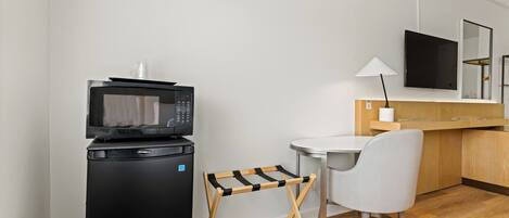 A compact kitchenette with a microwave, a small refrigerator, a wooden drying rack, a chair, and a table with a lamp.
