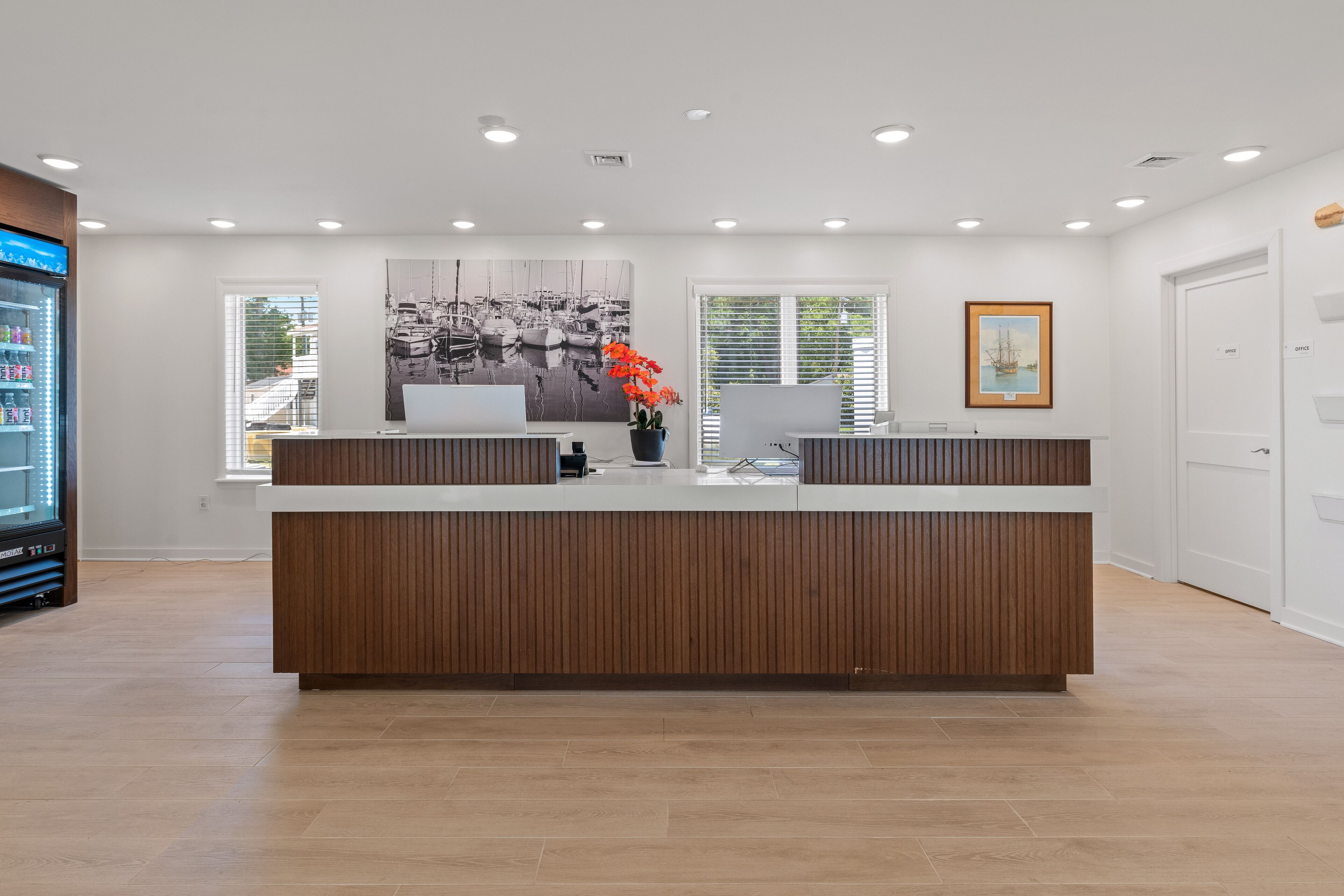 A modern reception desk with a wooden finish, a laptop, a vase with orange flowers, and a framed picture on the wall.