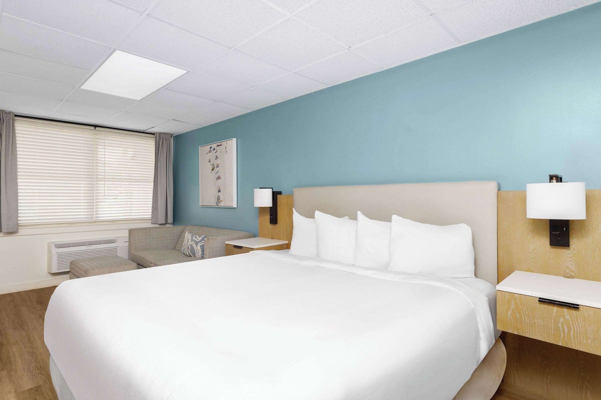 Habitación de hotel con una cama grande, una mesita de noche, una lámpara, una silla y una ventana con persianas.