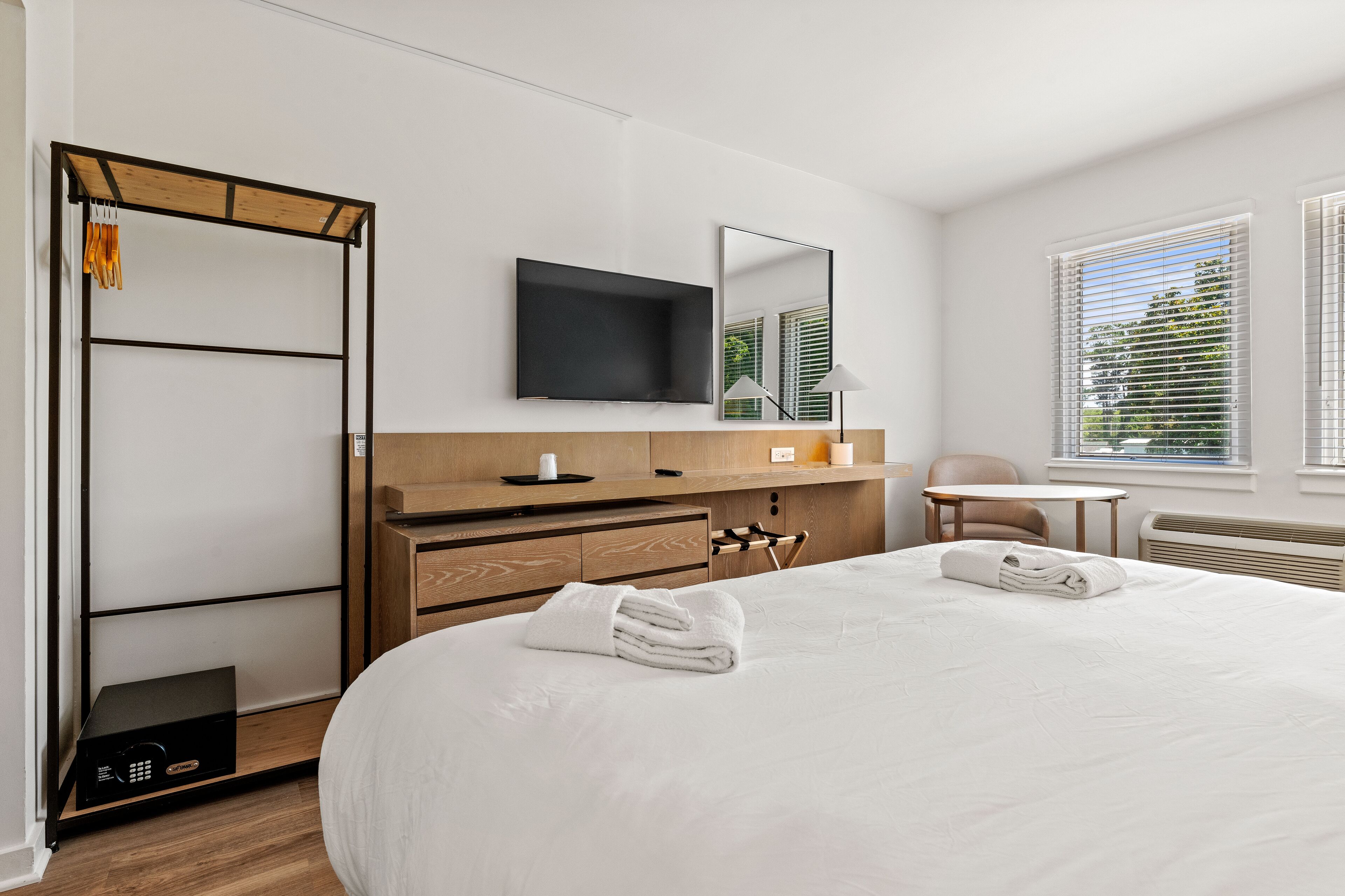 Une chambre d’hôtel avec un grand lit, une armoire en bois, un téléviseur à écran plat, un miroir, une petite table, une chaise et une fenêtre avec des stores.