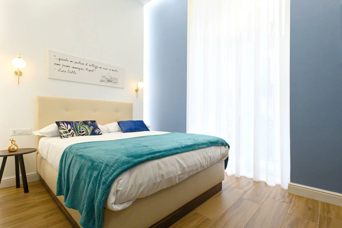 A neatly made bed with a teal blanket, a wall-mounted light fixture, and a framed quote on the wall.