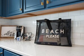 A kitchen counter with a coffee machine, a framed sign that reads 'coffee bar,' a black bag with 'BEACH PLEASE' text, and a coffee cup.