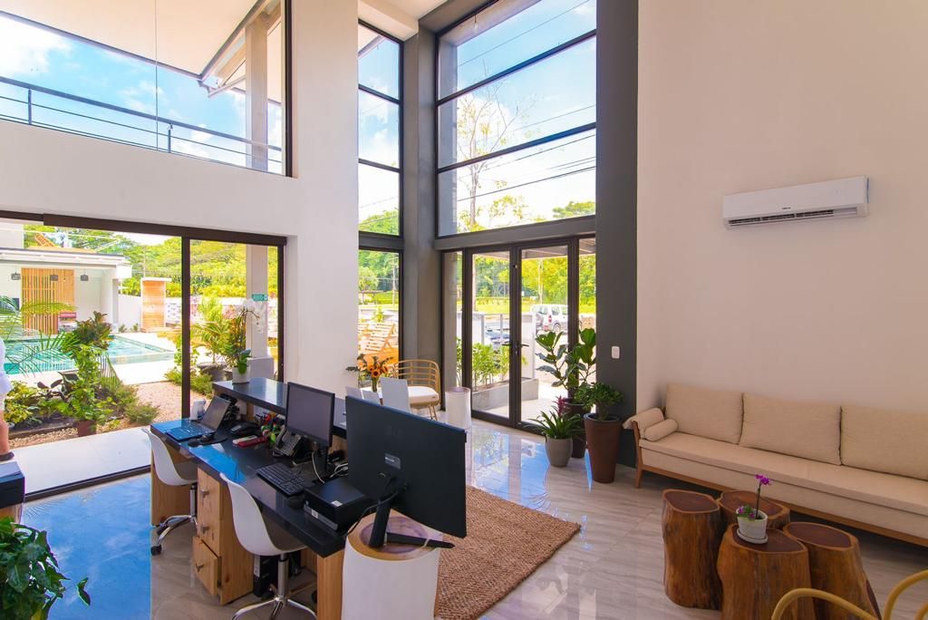 Un espacio de oficina moderno con ventanas amplias, un escritorio con computadoras, un sofá y una pequeña zona de piscina visible a través de las ventanas.