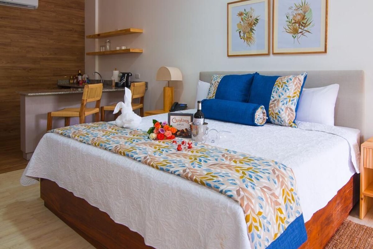 A hotel room with a bed, a desk, and a chair. There is a bottle of wine and a vase with flowers on the bed.