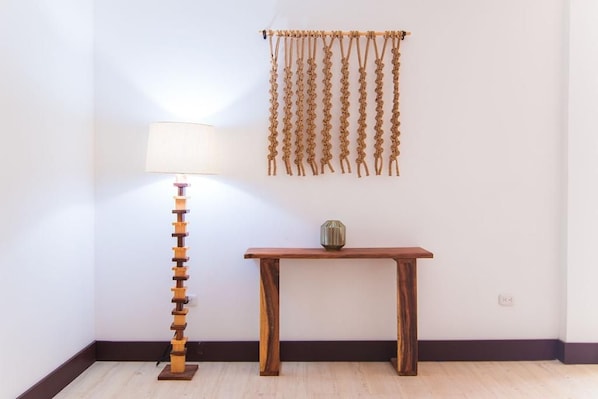 A corner of a room with a wooden floor, a lamp with a white shade, a wooden table with a vase, and a wall hanging.