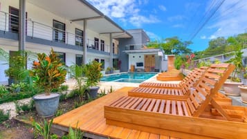 Un área de piscina moderna con tumbonas de madera y una piscina, rodeada de exuberante vegetación y un edificio de varios pisos.