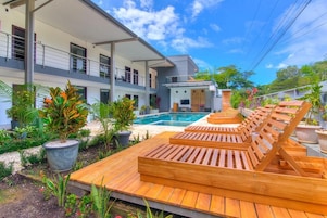 A modern poolside area with wooden loungers and a pool, surrounded by lush greenery and a multi-story building.