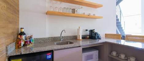 A compact kitchen with a built-in refrigerator, a sink, and a microwave. There are shelves above the sink holding glassware and a basket on the counter.