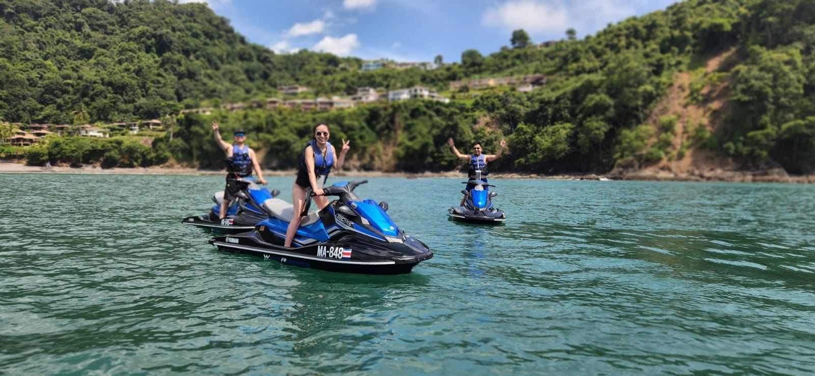 Three people on jet skis on a calm body of water with a forested hillside in the background.