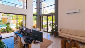 Espaço de escritório moderno com grandes janelas, uma mesa com computadores, um sofá e uma pequena área de piscina visível através das janelas.