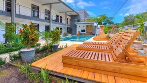 Uma área moderna à beira da piscina com espreguiçadeiras de madeira e piscina, rodeada por vegetação exuberante e um edifício de vários andares.