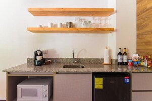 Cocina compacta con microondas, cafetera y refrigerador. Hay repisas de madera sobre el lavamanos con cristalería.