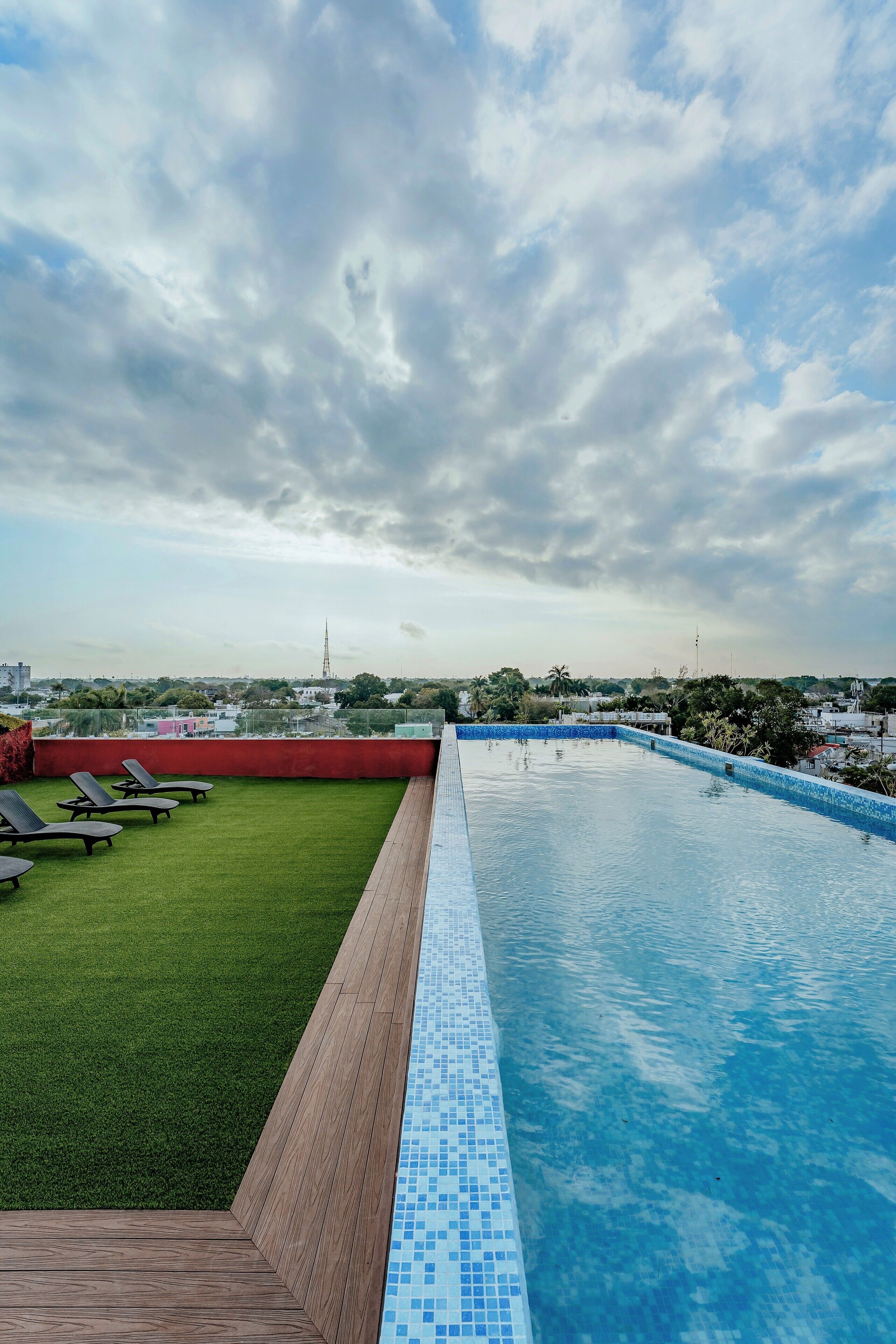 Una piscina en la azotea con vistas despejadas al horizonte urbano, rodeada de césped y sillones de descanso.