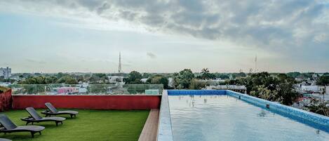 Una piscina en la azotea con vistas despejadas al horizonte urbano, rodeada de césped y sillones de descanso.
