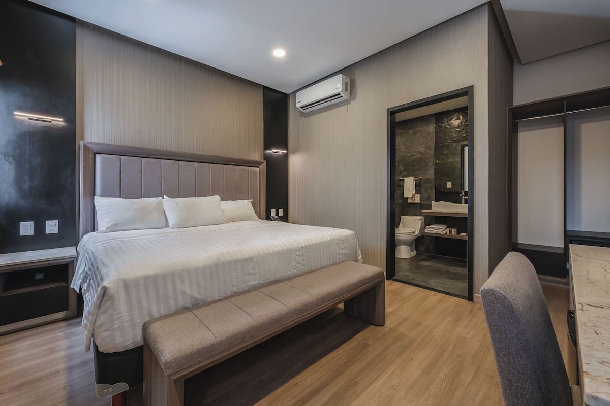 A modern hotel room with a large bed, a bench, a walk-in closet, and a bathroom visible through an open door.