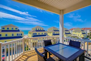 A covered outdoor area with a glass-top table and wicker chairs, overlooking a row of yellow buildings and a beach.