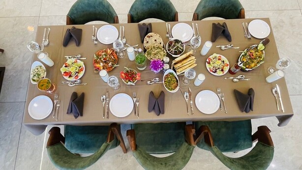 Mesa de comedor dispuesta con varios platos, cubiertos y diferentes fuentes, rodeada de sillas tapizadas en verde.