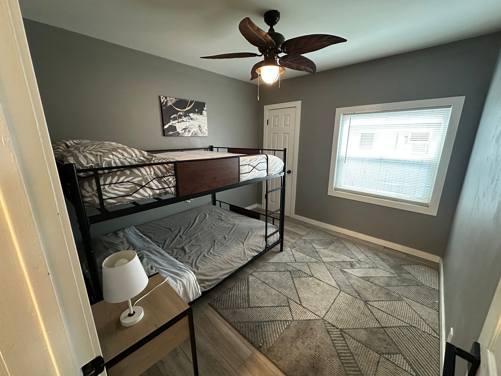 Ein kleines Schlafzimmer mit Etagenbett, Deckenventilator, Fenster mit Jalousien und einem Teppich mit geometrischem Muster.