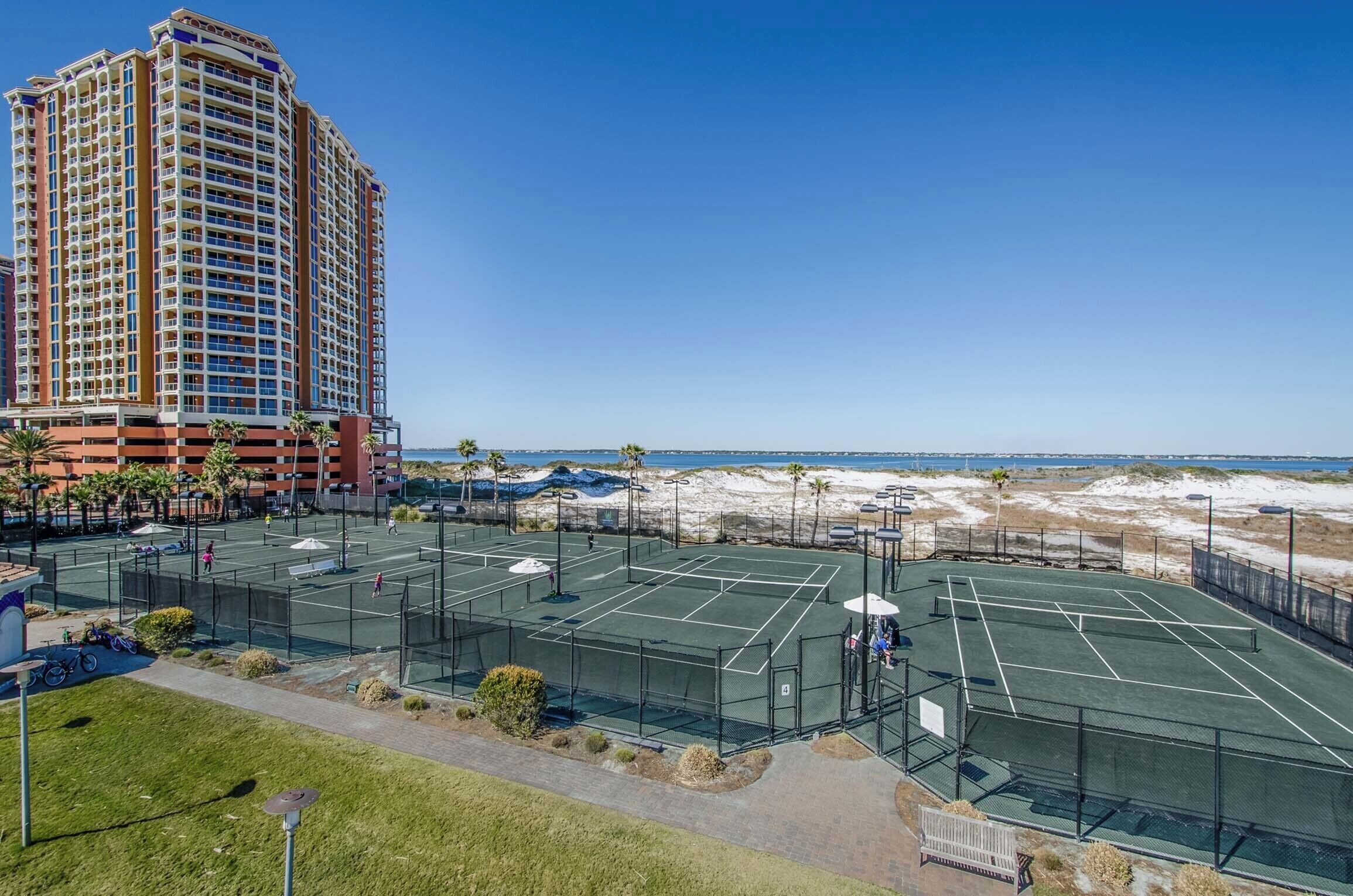 En tennisbana som ligger intill en strand, med ett högt hotell i bakgrunden.