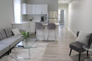 A modern living room with a sofa, two bar stools, a coffee table, and a kitchen area with white cabinets and a refrigerator. - WOW ★ Bright Apt ★ PRIME downtown Walk Everywhere (Albany)