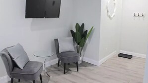 Ein modernes Zimmer mit einem Wand-Flachbildfernseher, zwei grauen Polsterstühlen, einem kleinen Glastisch, einer Topfpflanze und einer schwarzen Matte auf dem Boden.