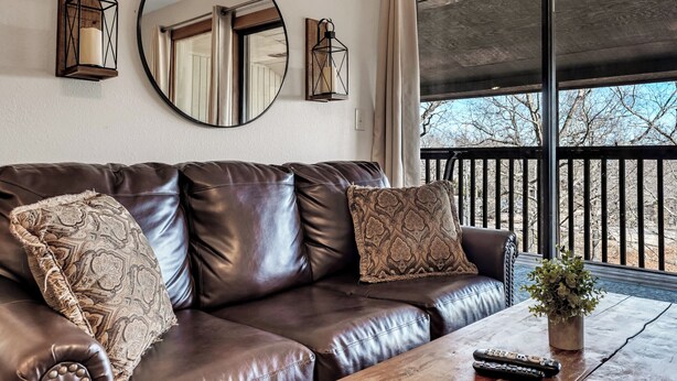 A brown leather sofa with patterned cushions, a wooden coffee table with a remote control, a mirror, and wall-mounted lanterns. - Gorgeous Branson Condo, VIEWS, Hike and Fish on Property, Private Lake, 2 Pools! (Reeds Spring)