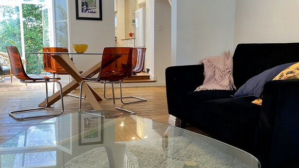 En moderne stue med et glasbord, en mørk sofa og et spiseområde med orange stole.