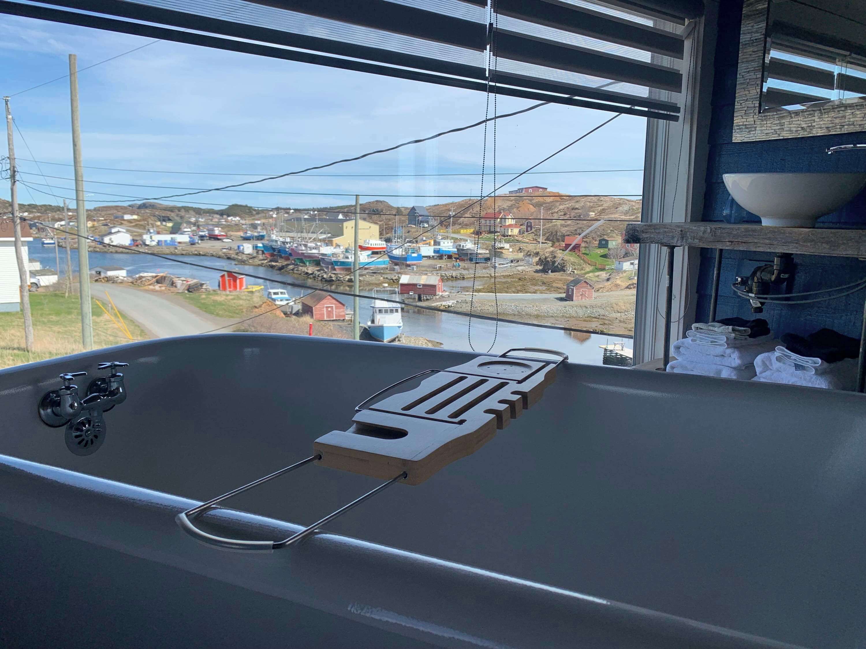 Une baignoire avec un plateau en bois, un lavabo avec un receveur, et une vue sur un village au bord de l’eau.