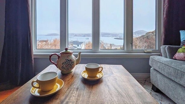 A room with a large window, a wooden table with two cups and a teapot, and a grey sofa. - On the Rocks Overlooking the Sea (Twillingate)