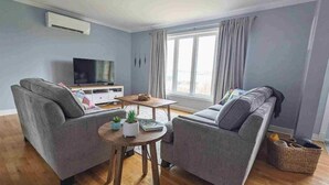 A living room with two sofas, a coffee table, a TV, and a large window with curtains. - On the Rocks Overlooking the Sea (Twillingate)