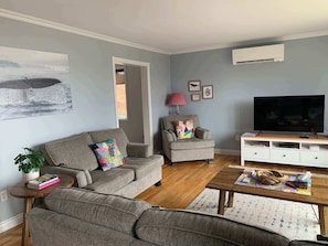 Ein Wohnzimmer mit grauem Sofa, einem kleinen Sessel, einem Couchtisch, einem Fernseher und einer weißen Unterhaltungsanlage.