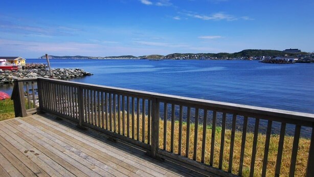 A wooden deck overlooking a calm body of water with distant hills and a clear sky. - Jagged Rocks (Twillingate)