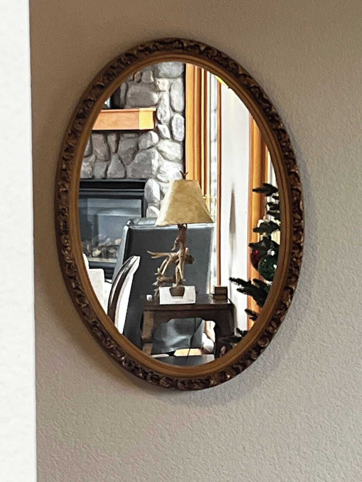 Un espejo con un marco decorativo refleja una habitación con chimenea de piedra, una mesa con lámpara y una silla.
