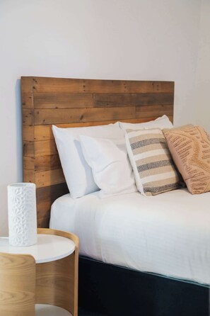 Un lit avec une literie blanche, une tête de lit en bois et une table de chevet sur laquelle se trouve un vase blanc.