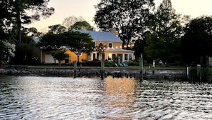 A house with a dock extending into a calm body of water, surrounded by trees and a stone wall. - Little piece of heaven in your own back yard! (New Bern)