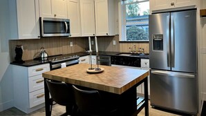 A modern kitchen with white cabinets, stainless steel appliances, a wooden island with chairs, and a window providing natural light. - Brand New Custom Built Cottage in Downtown w/Pool (Salem)