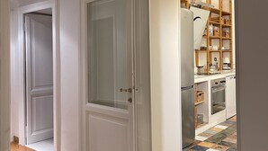 En hall med trägolv som leder till ett köksområde med kakel på väggen bakom arbetsbänken och hyllor i trä.