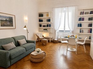 A living room with a green sofa, a coffee table, a dining table with chairs, and a bookshelf filled with books.